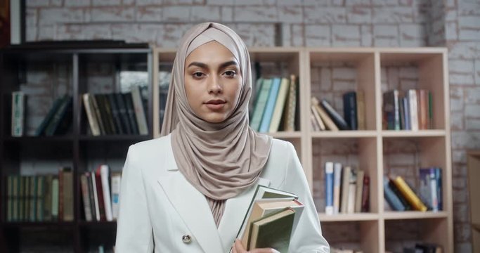 Female muslim student in library, holding books and looking at camera, girl wearing hijab spending time studying - modern islam, student lifestyle concept close up 4k - Powered by Adobe