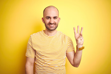 Naklejka premium Young bald man with beard wearing casual striped t-shirt over yellow isolated background showing and pointing up with fingers number three while smiling confident and happy.