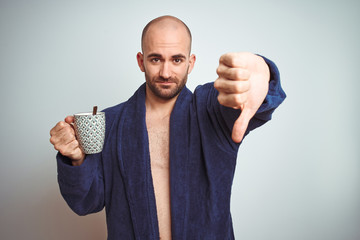 Naklejka premium Young man wearing a bathrobe drinking a cup of coffee in the morning over isolated background with angry face, negative sign showing dislike with thumbs down, rejection concept
