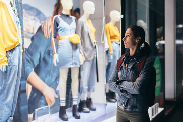 Girl stands outside the store and looks at the female clothes on the mannequins.