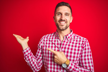 Naklejka premium Young handsome man over red isolated background amazed and smiling to the camera while presenting with hand and pointing with finger.