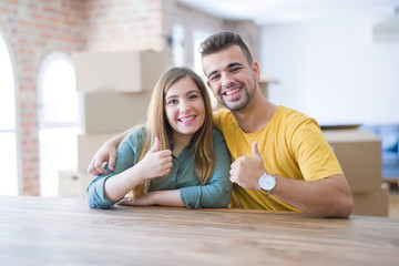 Young couple sitting on the table movinto to new home with carboard boxes behind them doing happy thumbs up gesture with hand. Approving expression looking at the camera with showing success.