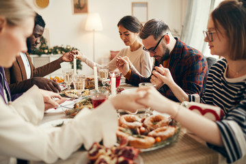 Multi-ethnic group of people sitting at dining table on Christmas and joining hands in prayer, copy...