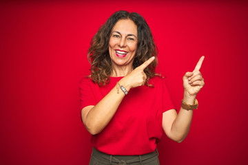 Middle age senior woman with curly hair over red isolated background smiling and looking at the camera pointing with two hands and fingers to the side.