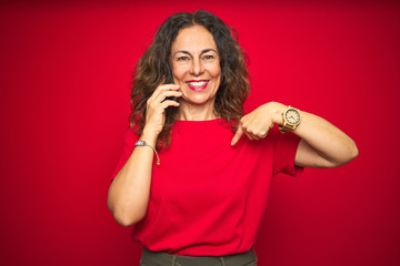 Middle age senior woman talking on the phone over red isolated background with surprise face pointing finger to himself