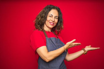 Middle age senior woman wearing apron uniform over red isolated background Inviting to enter smiling natural with open hand
