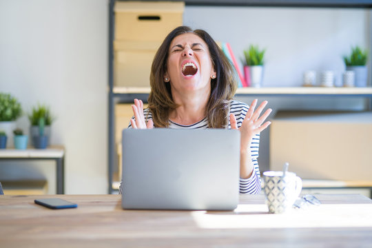 Middle age senior woman sitting at the table at home working using computer laptop crazy and mad shouting and yelling with aggressive expression and arms raised. Frustration concept.