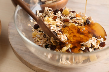 Pouring honey into bowl with oatmeal flakes and raisins on table, closeup. Healthy granola bar...