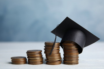 Coins and student graduation hat on white wooden table against blue background. Tuition fees concept