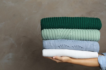 Woman holding stack of warm clothes on grey background, closeup. Autumn season