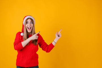 Fototapeta na wymiar excited woman pointing fingers at copy space wearing santa hat and red sweater isolated over yellow