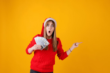 Fototapeta na wymiar happy girl dollar money in hand pointing at copyspace wearing santa hat isolated over yellow