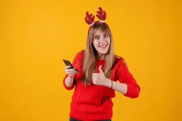 happy girl holding smartphone in hands wearing funny christmas hoop isolated over yellow