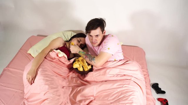 A young married couple are lying in bed, watching a film and eating chips. The husband feeds his wife chips. The woman is falling asleep from the boring movie.