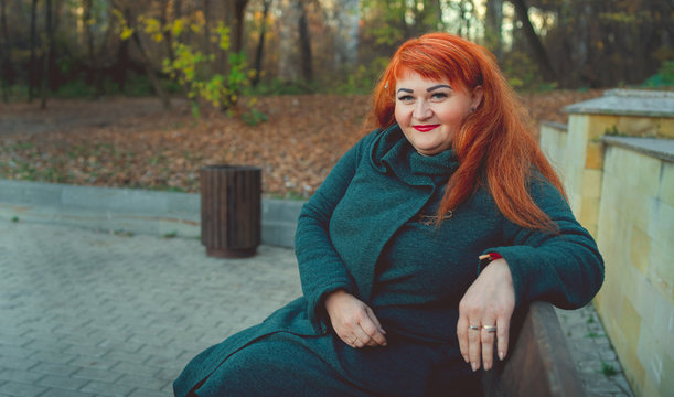 A portrait of a beautiful adult redhead woman, sitting on a bench near the vegetation.