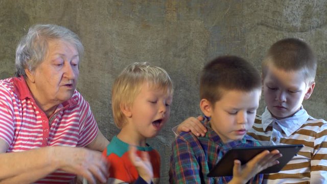 Three brothers in bright clothes play with tablet. Grandmother sits next to her grandsons and looks at their entertainment. Beautiful elderly woman with gray hair and wrinkles. Kids love granny