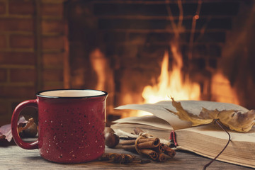 Red mug with hot tea and an open book in front of a burning fireplace, comfort, relaxation and warmth of the hearth concept