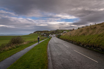 Young woman walking lonely along a road towards to a coastal city on a cold and rainy day with fields and the sea aside while grass is blown by the wind. Concept of being lonely and sadness.