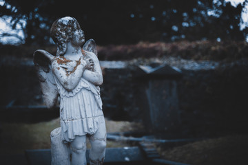 Obraz premium KILKENNY, IRELAND, DECEMBER 23, 2018: Sculpture of an old creepy cherub angel in the middle of a graveyard, full of lichen and mold, holding its chest and heart while looking at the cloudy sky.