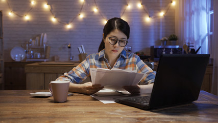 asian elegant woman in glasses at home dining desk using calculator and computer calculating budget. young japanese female reading document paper of payment in night kitchen. lady count house finance