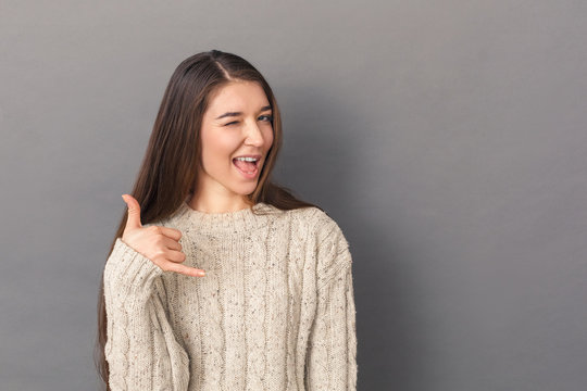 Freestyle. Young woman in sweater standing studio isolated on grey showing call gesture winking to camera playful
