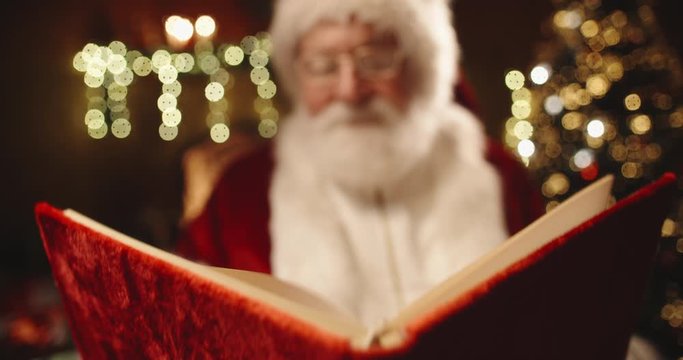 Bearded senior man in santa clause outfit flipping through pages of book with red cover in thematically decprated room - christmas spirit concept close up 4k footage