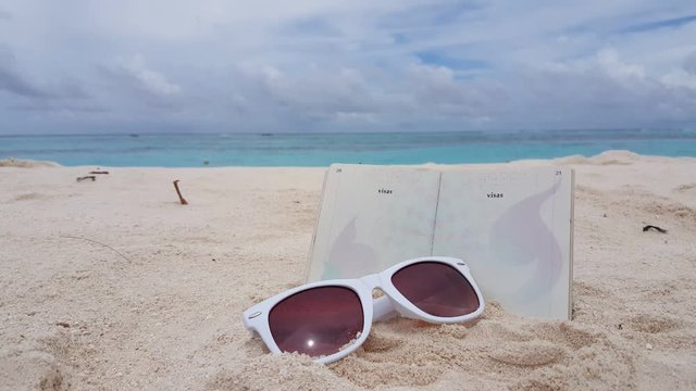 Copy space with sunglasses and passport plunged on white beach of tropical island with sea and cloudy sky background