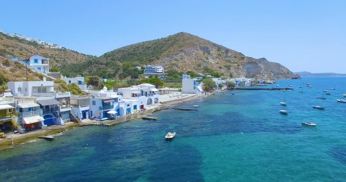 Circling aerial above beautiful vintage fishing village with boats floating in crystal clear water on the island of Milos Greece in the Aegean Sea of the Mediterranean. Daytime exterior landscape.