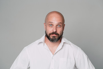 Obraz premium Handsome serious middle-aged man in a white shirt looks at the camera in surprise and holds his hands near his face on a gray background