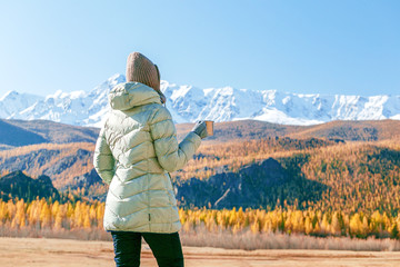  A young tourist woman drinks a hot drink from a cup and enjoys the scenery in the autumn mountains. Trekking concept
