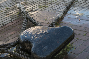 A wet mooring bollard painted black, with three thick braided mooring ropes of moored vessels attached to it, mounted on a wharf covered with gray and brown paving slabs with puddles on them