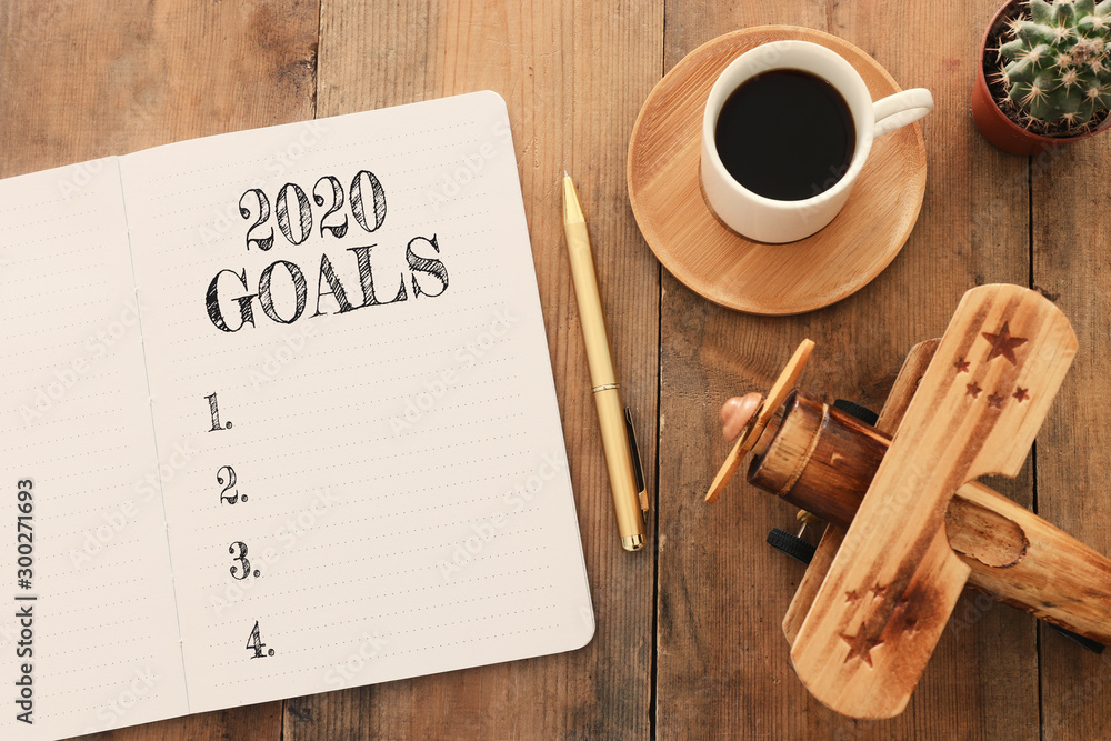 Wall mural Business concept of top view 2020 goals list with notebook, cup of coffee and old plane toy over wooden desk