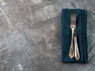 Silver fork and knife on dark green emerald linen napkin over gray textured background table with...
