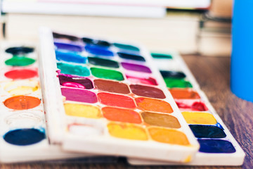 Watercolor paint, brushes well used on wooden table with stack of books background.
