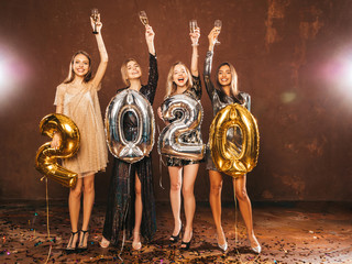 Beautiful Women Celebrating New Year.Happy Gorgeous Girls In Stylish Sexy Party Dresses Holding Gold and Silver 2020 Balloons, Having Fun At New Year's Eve Party.Сarrying and drinking champagne flutes