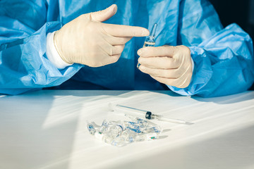 doctor in a blue surgical gown and mask holds in his hand a medical syringe and bottle with insulin for diabetes. Many drugs for injection on white table.