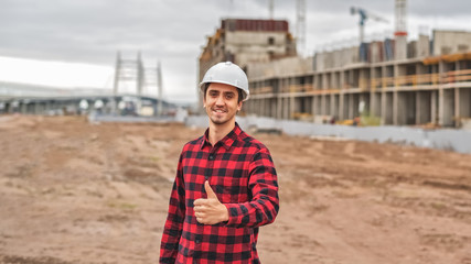 civil engineer in a white helmet on the background of construction, with a raised thumb.