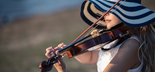 Violinist woman wearing white dress with hat playing violin at lakeside