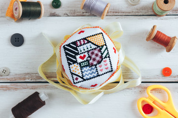 Handmade and needlework. Handmade pin cushion with a simple ornament and a pair of scissors on a white table. Sewing supplies in the background. Top view