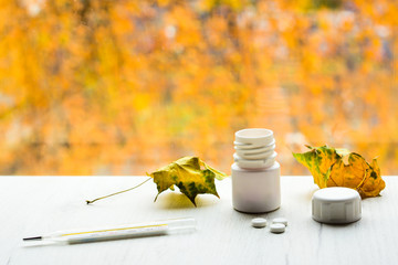 Thermometer, tablets from white bottle and yellow leaves on white table, with autumn colors on background. Cold and flu season treatment concept.