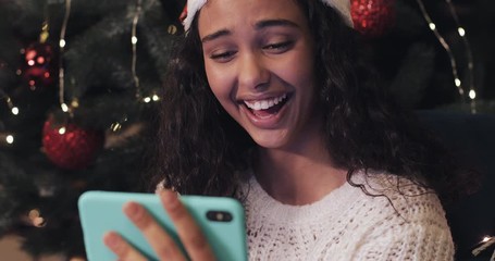Happy Beautiful Girl in Santas Hat Sitting at Christmas Tree at the Background, Using her Smartphone, Looking at the Screen and Laughing. Holiday Concept. Close Up - Powered by Adobe