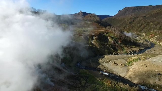 pictorial valley of geysers with white steam over green and brown hilly canyon landscape aerial view