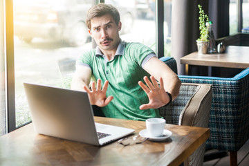 Fototapeta premium No, stop, please don't touch me! Young scared businessman in green t-shirt sitting with laptop, looking with afraid face and stop sign. business problem concept. indoor shot near big window at daytime