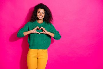 Portrait of positive dreamy afro american girl show heart from fingers symbol of her love to boyfriend on valentine day in winter wear knitted pullover yellow pants isolated pink color background