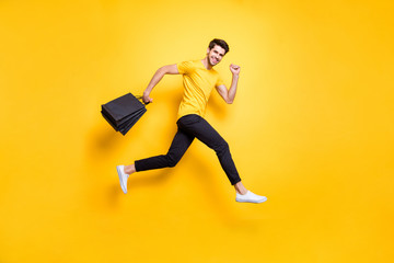 Fototapeta na wymiar Full size photo of handsome guy jumping high holding many packages speed rushing sale shopping wear casual t-shirt black pants isolated yellow color background