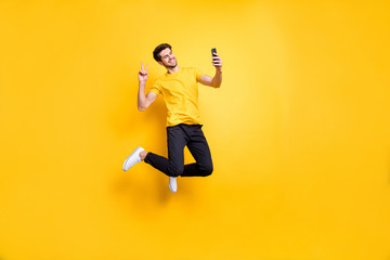 Fototapeta na wymiar Full length photo of handsome guy jumping high holding telephone taking selfies showing v-sign symbol wear casual t-shirt pants isolated yellow color background