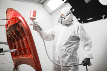 Auto mechanic worker painting red car hood with spray gun in a paint chamber during repair work.