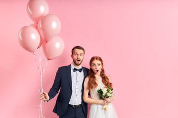 Surprised couple in wedding dress and tuxedo stand together isolated over pink background. With...