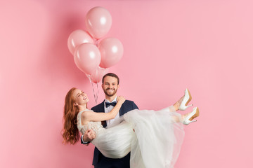Attractive redhaired woman wearing white wedding dress sit on man's hands. Cheerful smiling couple...