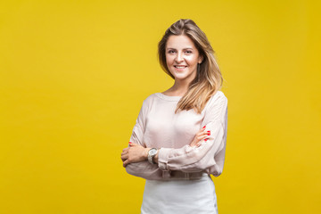 Portrait of happy attractive young woman with fair hair in casual beige blouse standing with crossed arms and looking at camera with toothy smile. indoor studio shot isolated on yellow background